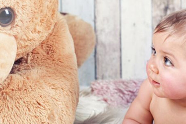 Baby schaut liegend großen Teddybären an